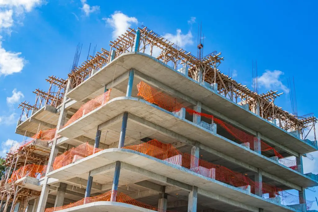 Multi-story concrete building under construction with orange safety netting and wooden scaffolding against a blue sky.