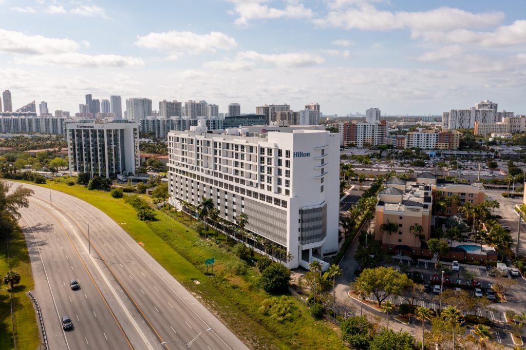 Aerial view of a coastal city with a prominent white Hilton hotel in the foreground, tall apartment blocks in the distance, and a multi-lane highway curves along the left edge.