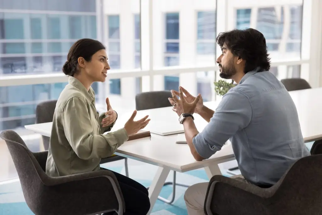 Two coworkers sit across from each other at a white table, gesturing with their hands during a discussion in a bright office setting.