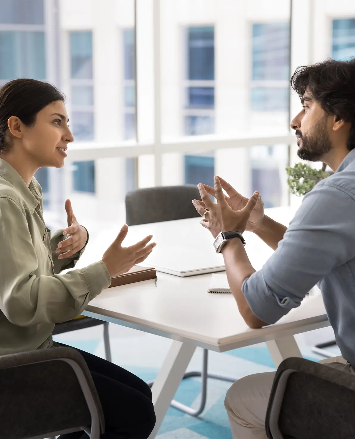 Two colleagues seated at a white table in a bright office, gesturing with their hands as they talk.
